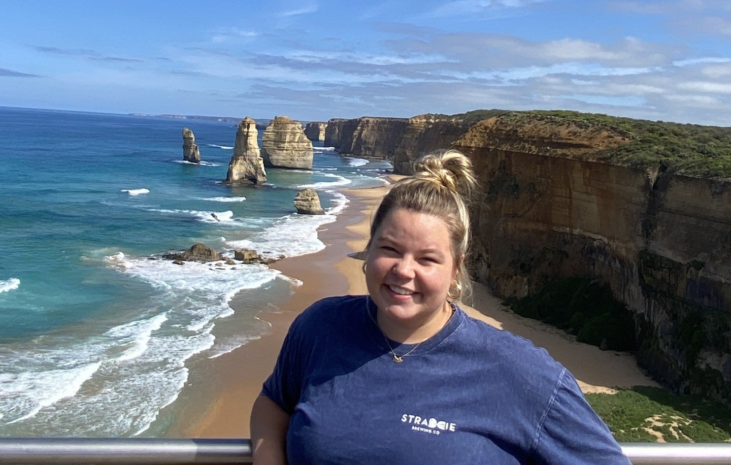 A white female with blond hair pulled back wearing a blue T-shirt stands on a cliff overlooking a sandy beach with turquoise waves, rocky formations, and coastal cliffs below.
