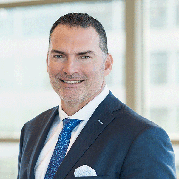 A smiling person in a blue suit and patterned tie, standing indoors with a softly blurred background.