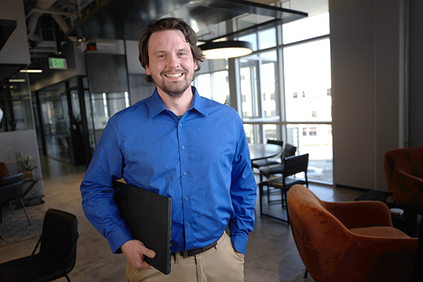 Man in a blue button-down shirt standing in a modern office lounge, holding a laptop and smiling toward the camera.