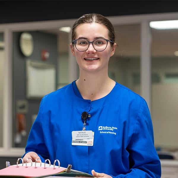 A person wearing a bright blue Indiana State University nursing uniform and a name badge, holding a pink binder while standing in a clinical setting with medical equipment and a wall clock in the background.
