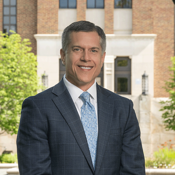 A smiling, middle-aged man with dark hair, and wearing a dark suit and light blue tie, stands outdoors in front of a brick academic building with columns and framed by greenery.