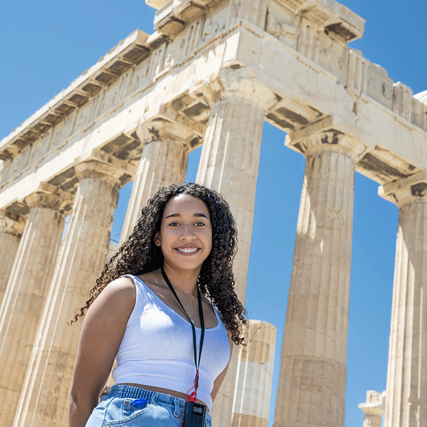 A smiling young woman stands in front of the Parthenon in Athens, wearing a white sleeveless top and blue jeans, with tall stone columns rising behind her against a clear blue sky.