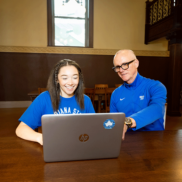 A student and a financial aid counselor sit at a wooden table indoors, working together on an HP laptop, with the counselor pointing at the screen; both wear Indiana State University apparel, and the room features warm lighting, wood furniture, and a window in the background.