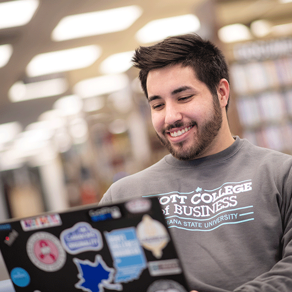 A smiling man with dark hair and a beard stands in a library holding an open laptop covered with stickers, and wearing a grey weatshirt that reads ‘Scott College of Business, Indiana State University,’ with bookshelves softly visible in the background.