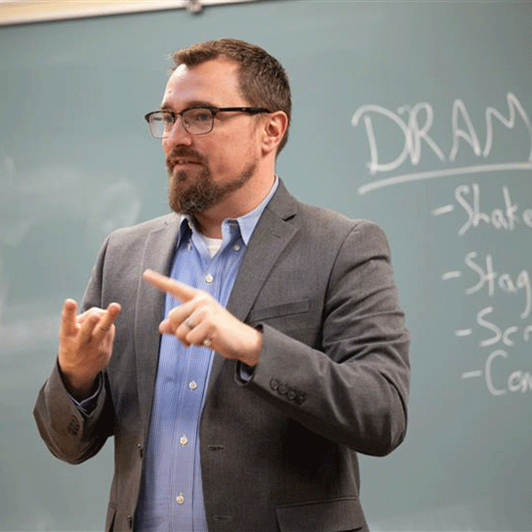 A male instructor wearing glasses and a grey sport jacket over a blue shirt stands in front of a classroom chalkboard, gesturing while explaining a lesson; the board displays the heading ‘DRAM’ with several bullet points written beneath it.
