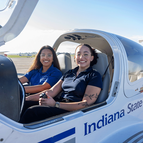 Two women seated inside a small Indiana State University training aircraft on a runway, viewed through the open cockpit door, with the plane’s interior controls visible and a clear sky in the background.