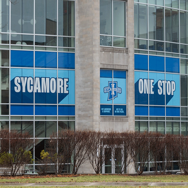 Exterior of a modern glass and brick campus building with large blue window graphics reading ‘Sycamore One Stop,’ trees and landscaping visible along the front.