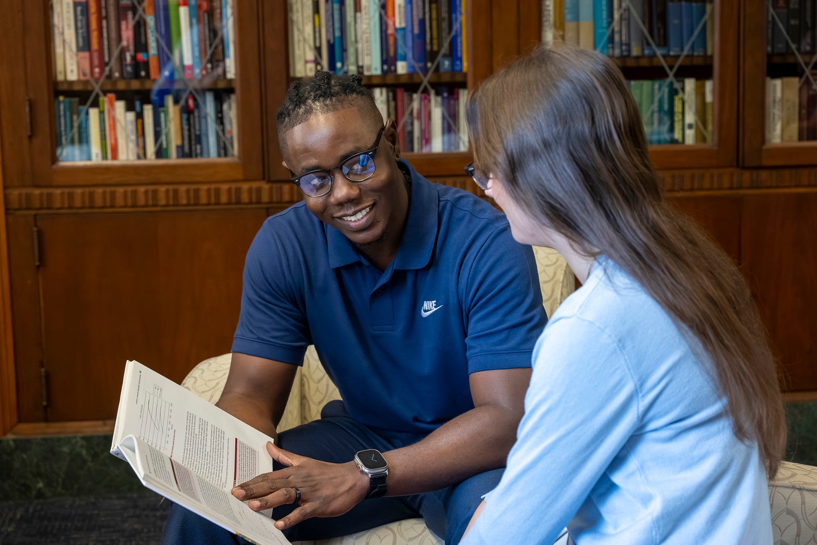 Student and tutor sitting together in a library, reviewing an open textbook and discussing the material in front of bookshelves.