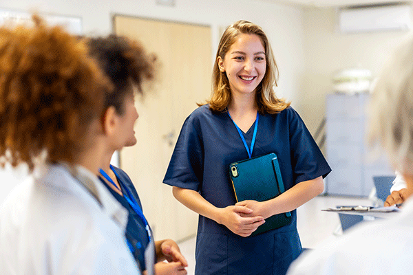 Smiling healthcare professional in navy scrubs holding a tablet and speaking with colleagues in a bright clinical setting.