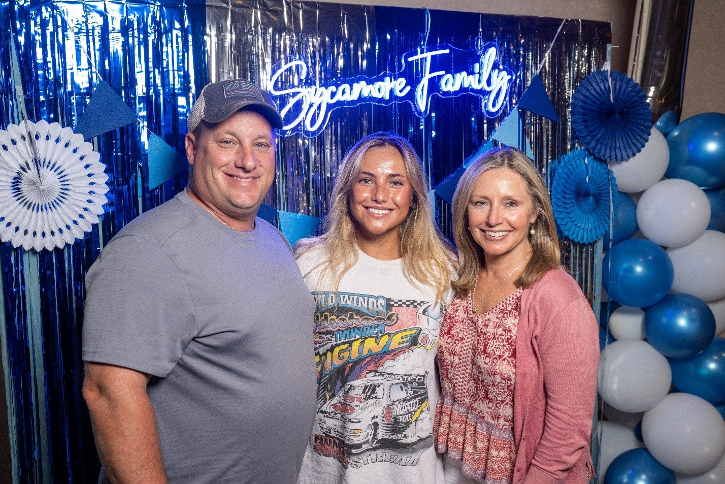 Three people—two adults and a young woman—stand smiling together in front of a blue party backdrop with metallic streamers, balloons, and a glowing sign that reads “Sycamore Family.”