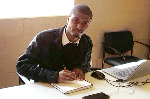 A Black male student is seated at a desk indoors, writing in a notebook beside an open laptop, with a phone and computer mouse on the table and empty chairs against the wall nearby.