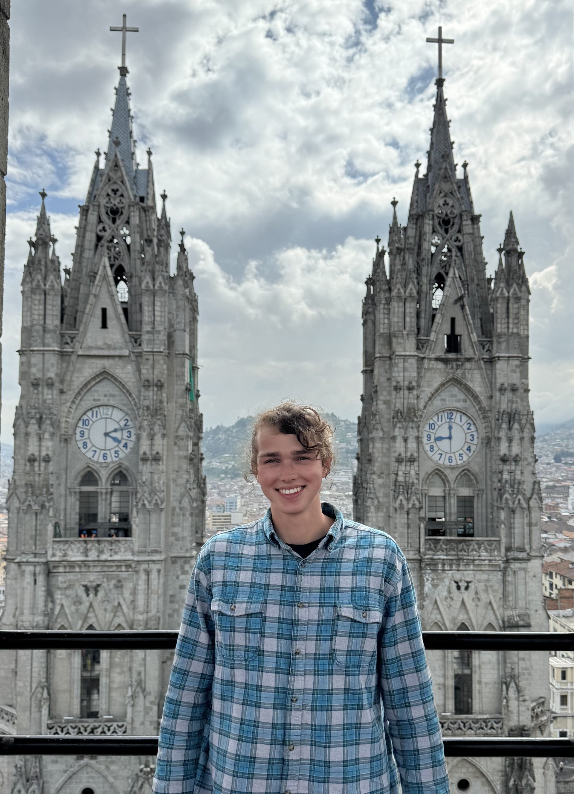 Young man smiling in front of two tall Gothic-style cathedral towers with large clocks, under a cloudy sky, overlooking a city.