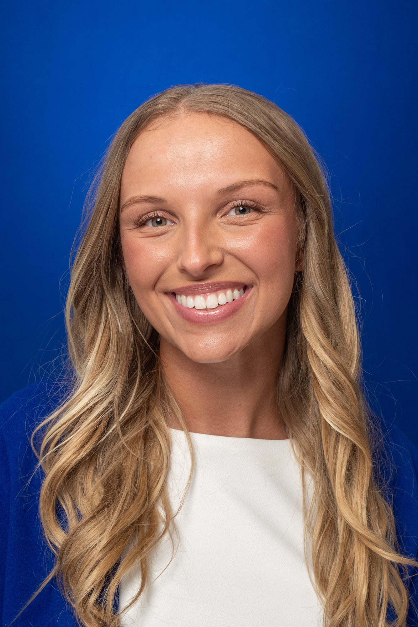 The image is a professional headshot of a smiling person against a blue studio background. They have long wavy blonde hair and are wearing a bright blue blazer over a white top. The lighting and composition are polished and consistent with a corporate, academic, or conference profile photo.