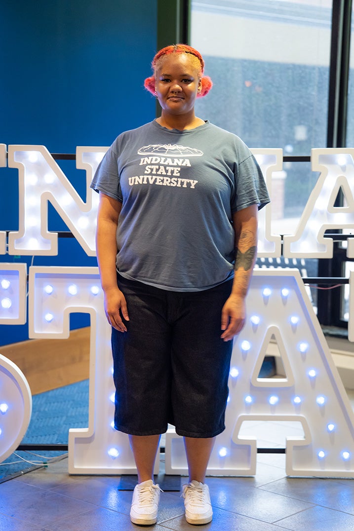 A person with bright pink braided hair stands indoors in front of illuminated “Indiana State” display letters. They are wearing a blue Indiana State University T-shirt, black knee-length shorts, and white sneakers. A tattoo is visible on one forearm, and large windows and blue walls are in the background.