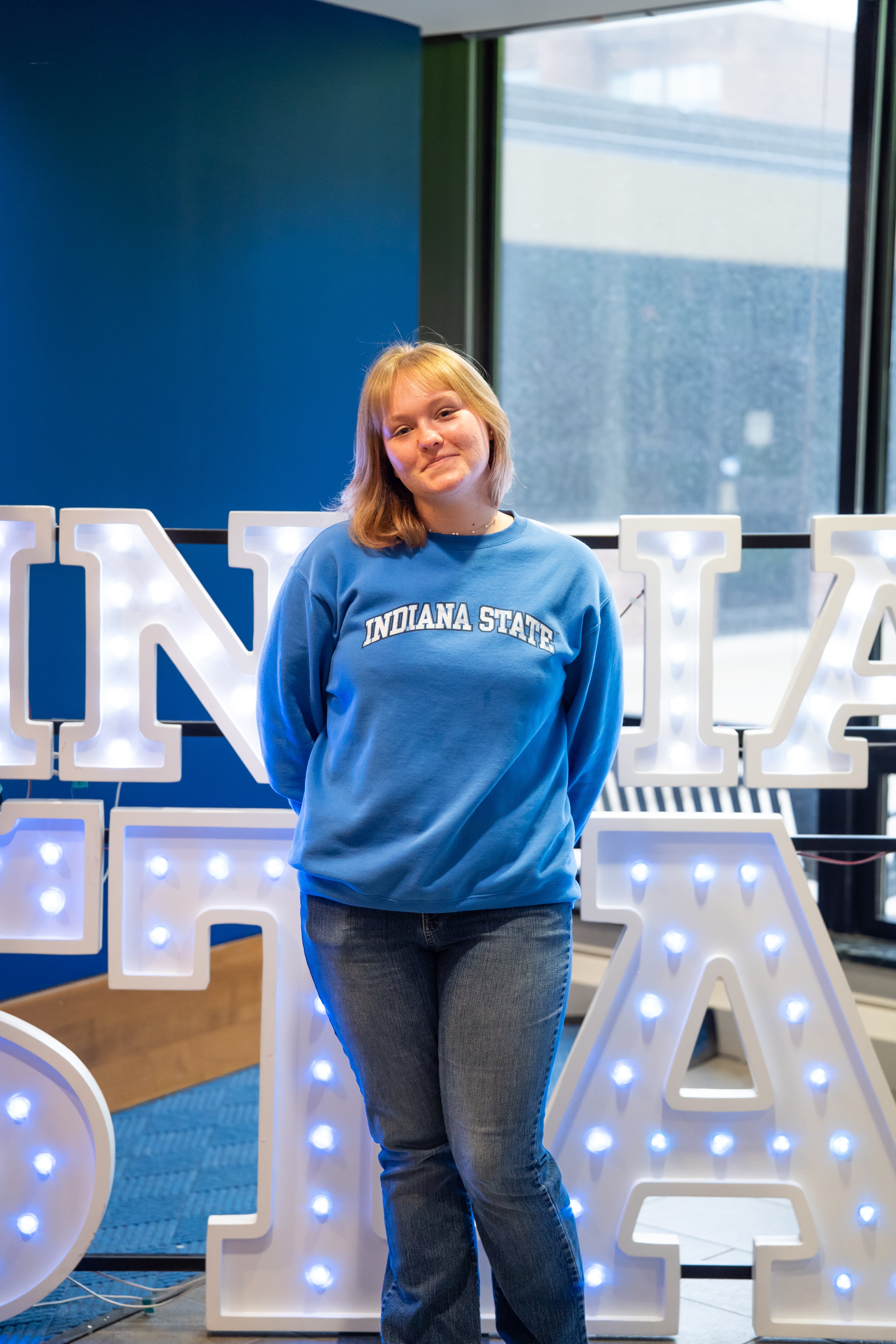 A person with short blonde hair stands indoors in front of illuminated “Indiana State” display letters and large windows. They are wearing a bright blue Indiana State sweatshirt and dark blue jeans, with their hands behind their back and a slight smile. Blue walls and lit signage are visible in the background.