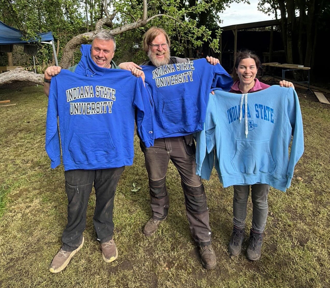 Three people standing outdoors, each holding up a blue Indiana State University hoodie. They are smiling in a grassy area with trees and a building in the background.