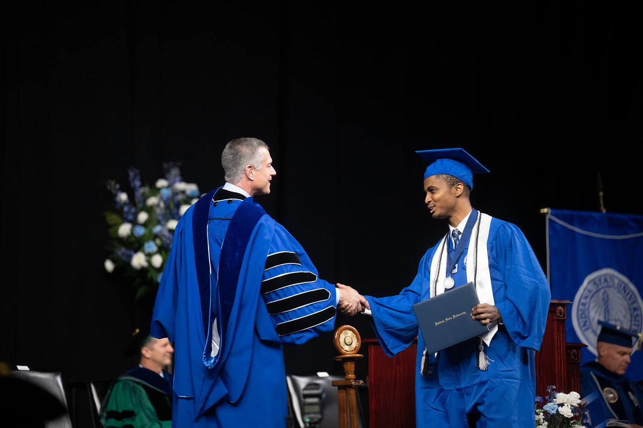Young Black male in a blue cap and gown shakes hands with a Indiana State University President Dr. Mike Godard on stage while holding a diploma. The graduate wears a white stole, honor cords, and a medal. A podium, seated faculty, and stage decorations are visible in the background.