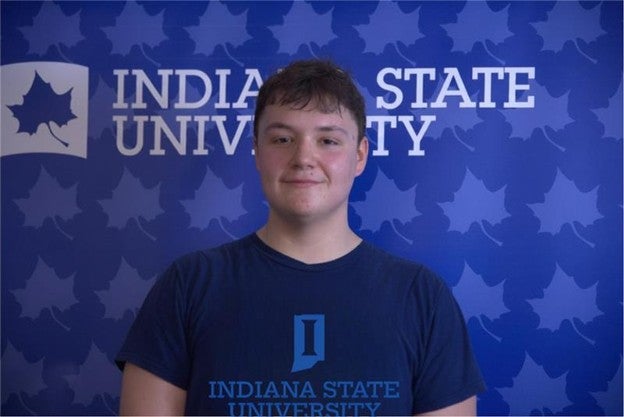 A person with short brown hair stands in front of a blue Indiana State University backdrop patterned with leaf logos. They are wearing a navy blue Indiana State University T-shirt and smiling slightly at the camera.