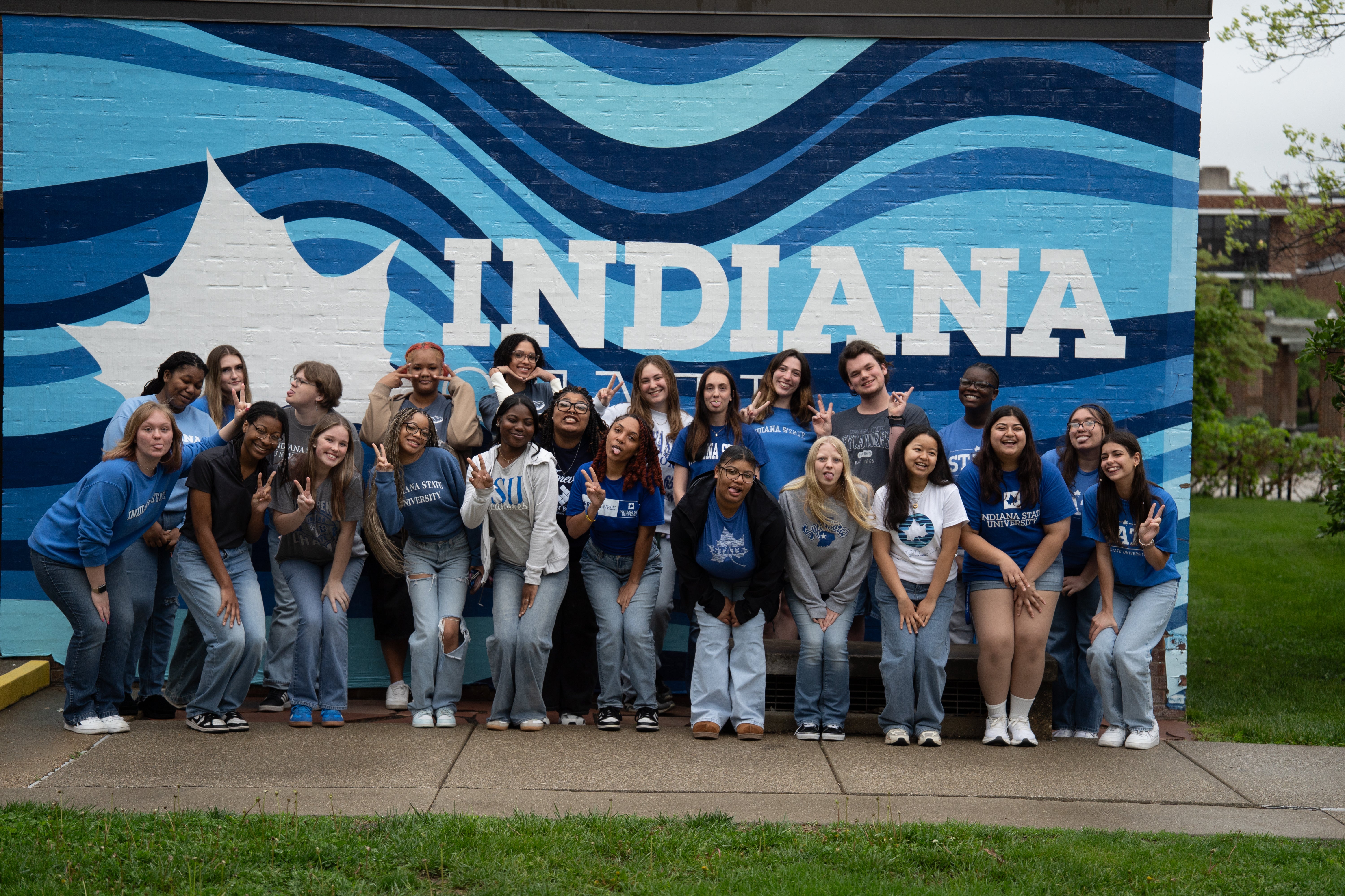 A large group of students poses together outdoors in front of a blue Indiana State mural wall with large white lettering. Many of the students are smiling, making peace signs, or striking playful poses while wearing Indiana State clothing in shades of blue, white, and gray. Grass, sidewalks, and campus buildings are visible around the mural.
