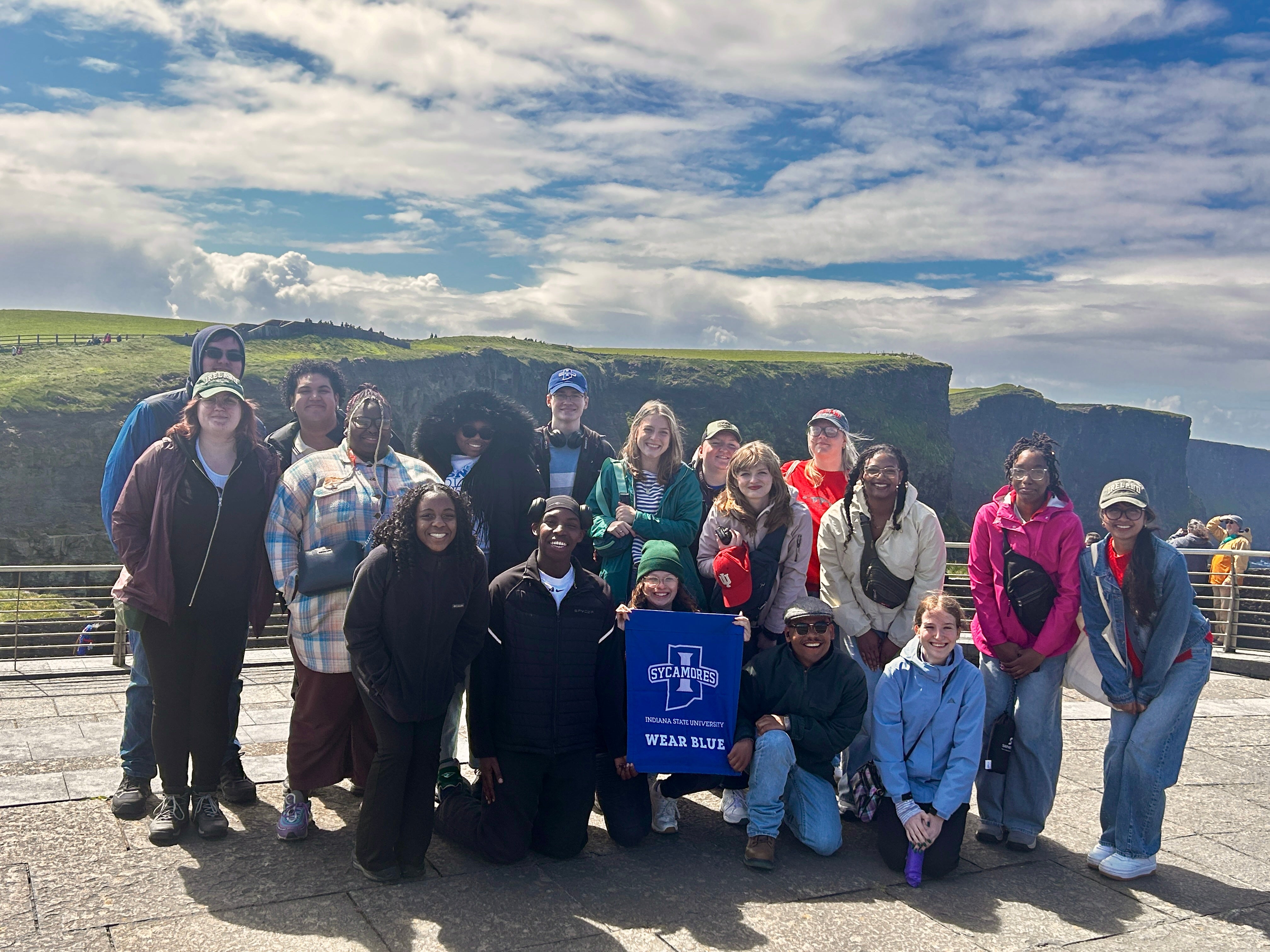 A group of Indiana State Students smiling in front of the Cliffs of Moher in Ireland. One individual is holding a Indiana State University sign. The sky is partly cloudy, and the landscape features green cliffs and ocean views in the background.