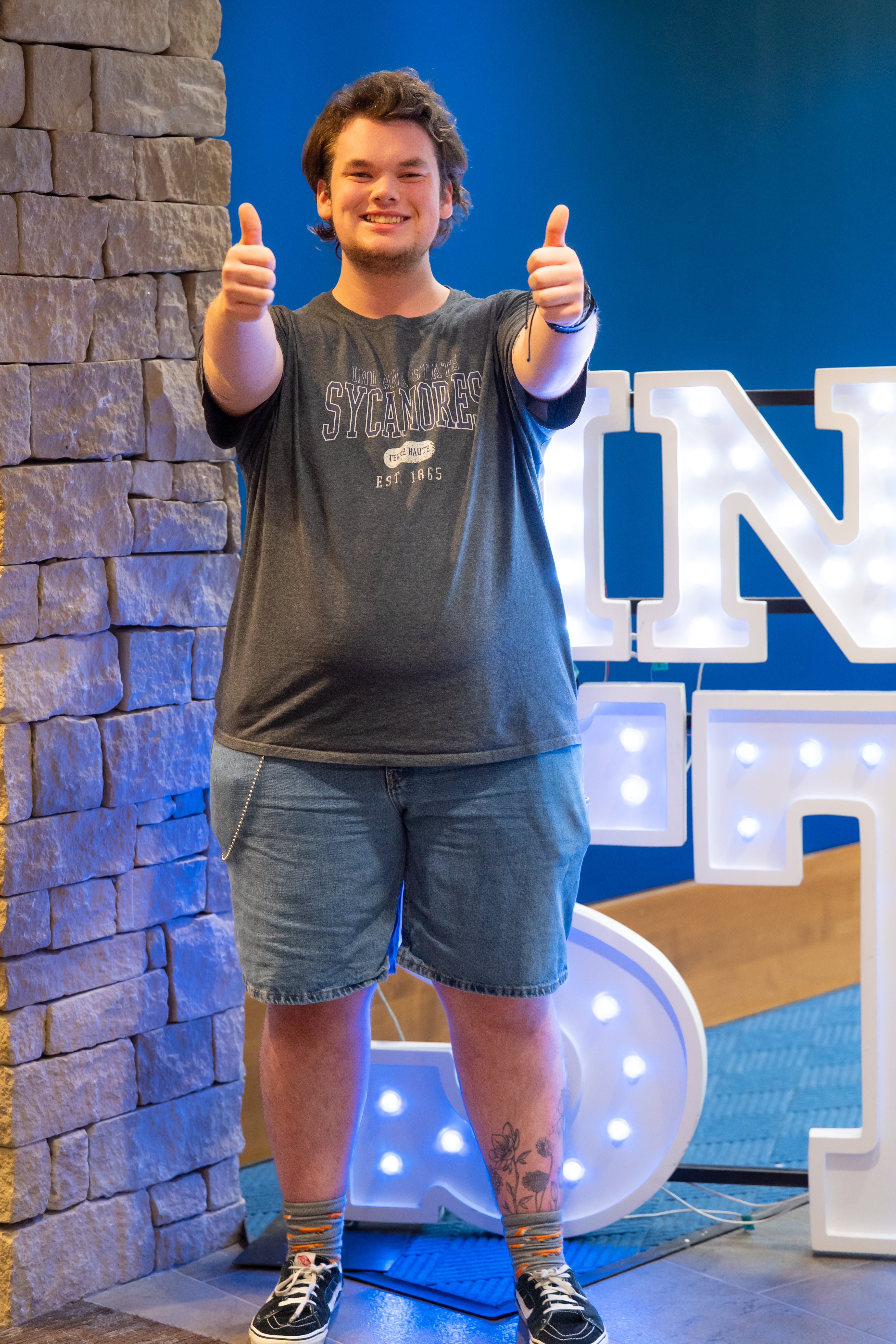 A smiling person with medium-length dark hair stands indoors beside a stone wall and illuminated “Indiana State” display letters, giving two thumbs up toward the camera. They are wearing a dark gray Indiana State Sycamores T-shirt, denim shorts, patterned socks, and black sneakers, with a floral tattoo visible on one lower leg. Blue walls and lit signage are visible in the background.