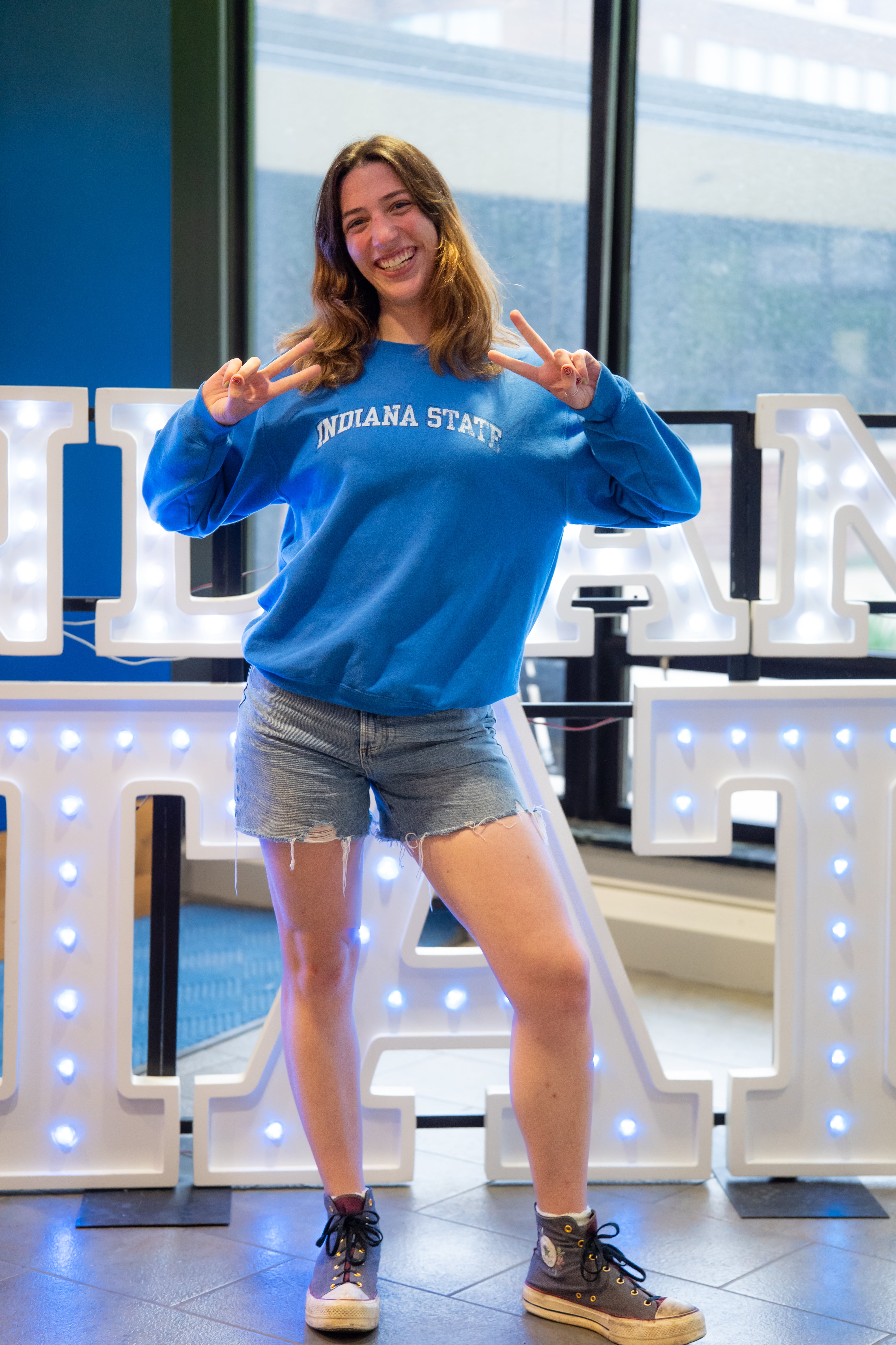 A smiling person with shoulder-length brown hair poses indoors in front of illuminated “Indiana State” display letters and large windows, holding up peace signs with both hands. They are wearing a bright blue Indiana State sweatshirt, denim shorts, and dark high-top sneakers. Blue walls and lit signage are visible in the background.