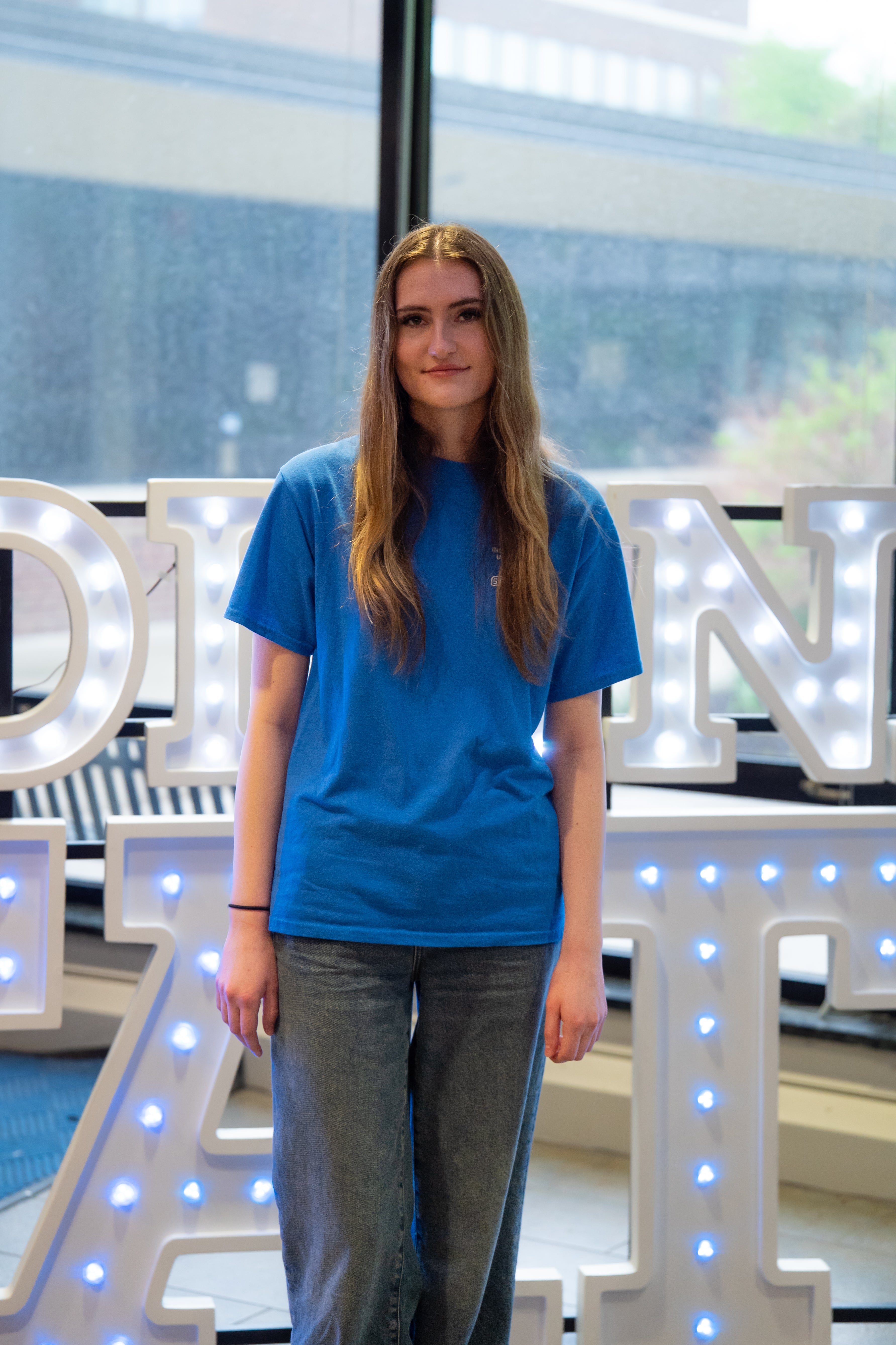 A person with long light brown hair stands indoors in front of illuminated “Indiana State” display letters and large windows. They are wearing a bright blue T-shirt and blue jeans, with their arms relaxed at their sides and a neutral expression. Blue walls and lit signage are visible in the background.