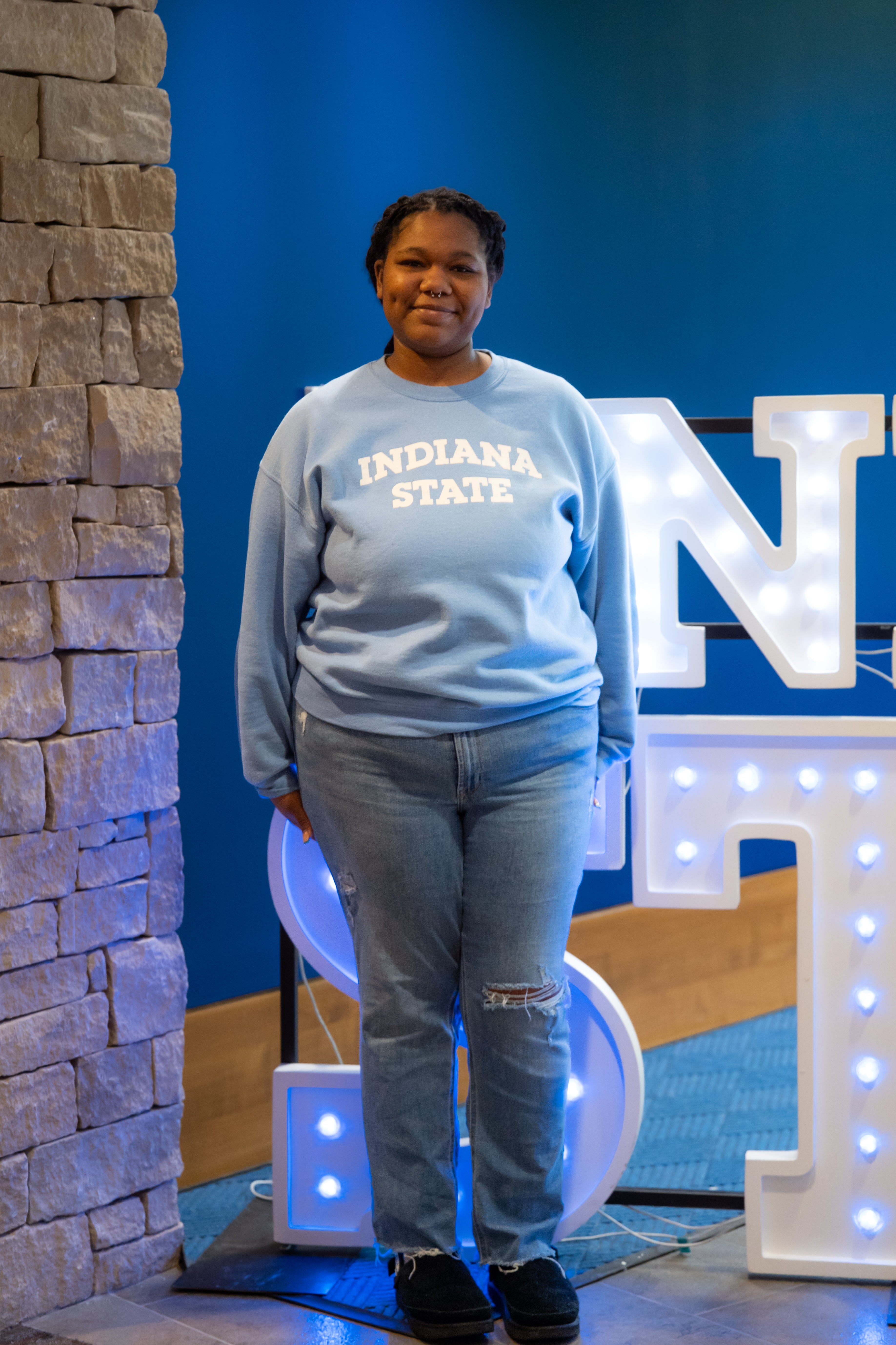 A smiling person with braided hair and a septum piercing stands indoors beside a stone wall and illuminated “Indiana State” display letters. They are wearing a light blue Indiana State sweatshirt, ripped light blue jeans, and black shoes, with their hands relaxed at their sides. Blue walls and lit signage are visible in the background.