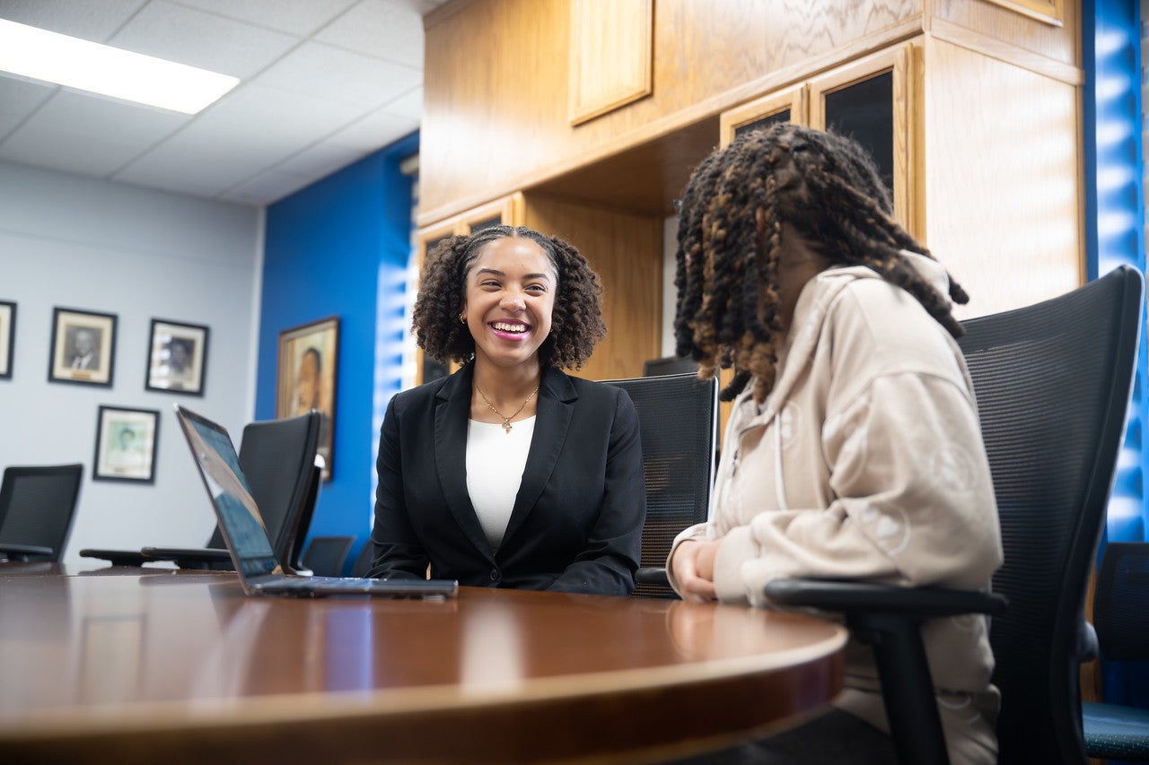 Two people are seated at a conference table in an office or meeting room. One person in a black blazer is smiling and appears engaged in conversation with the other person, who is turned slightly away from the camera. A laptop is open on the table, and the room has wood cabinetry, framed photos on the walls, and blue accent walls. The atmosphere appears professional and friendly.