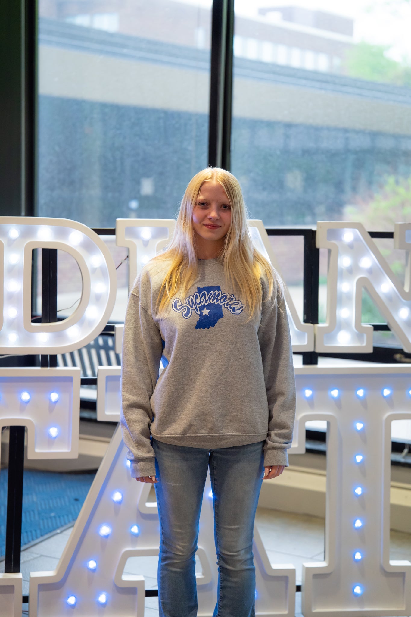 A person with long blonde hair stands indoors in front of illuminated “Indiana State” display letters and large windows. They are wearing a gray Sycamores sweatshirt and light blue jeans, with their arms relaxed at their sides and a neutral expression. Blue walls and lit signage are visible in the background.