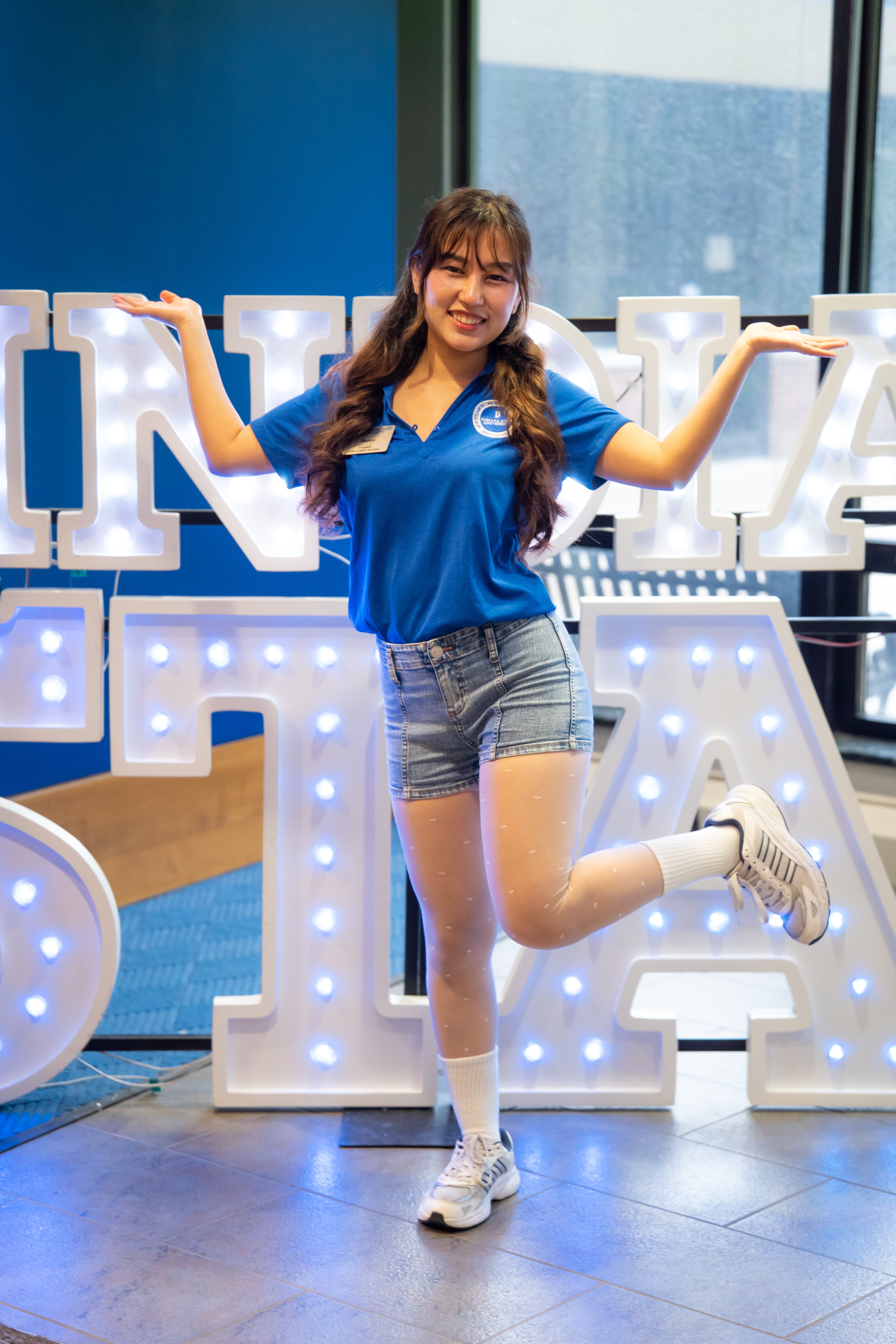 A smiling person with long dark wavy hair poses indoors in front of illuminated “Indiana State” display letters and large windows, balancing on one leg with both arms raised outward. They are wearing a bright blue collared shirt with a name tag, denim shorts, white socks, and white sneakers. Blue walls and lit signage are visible in the background.