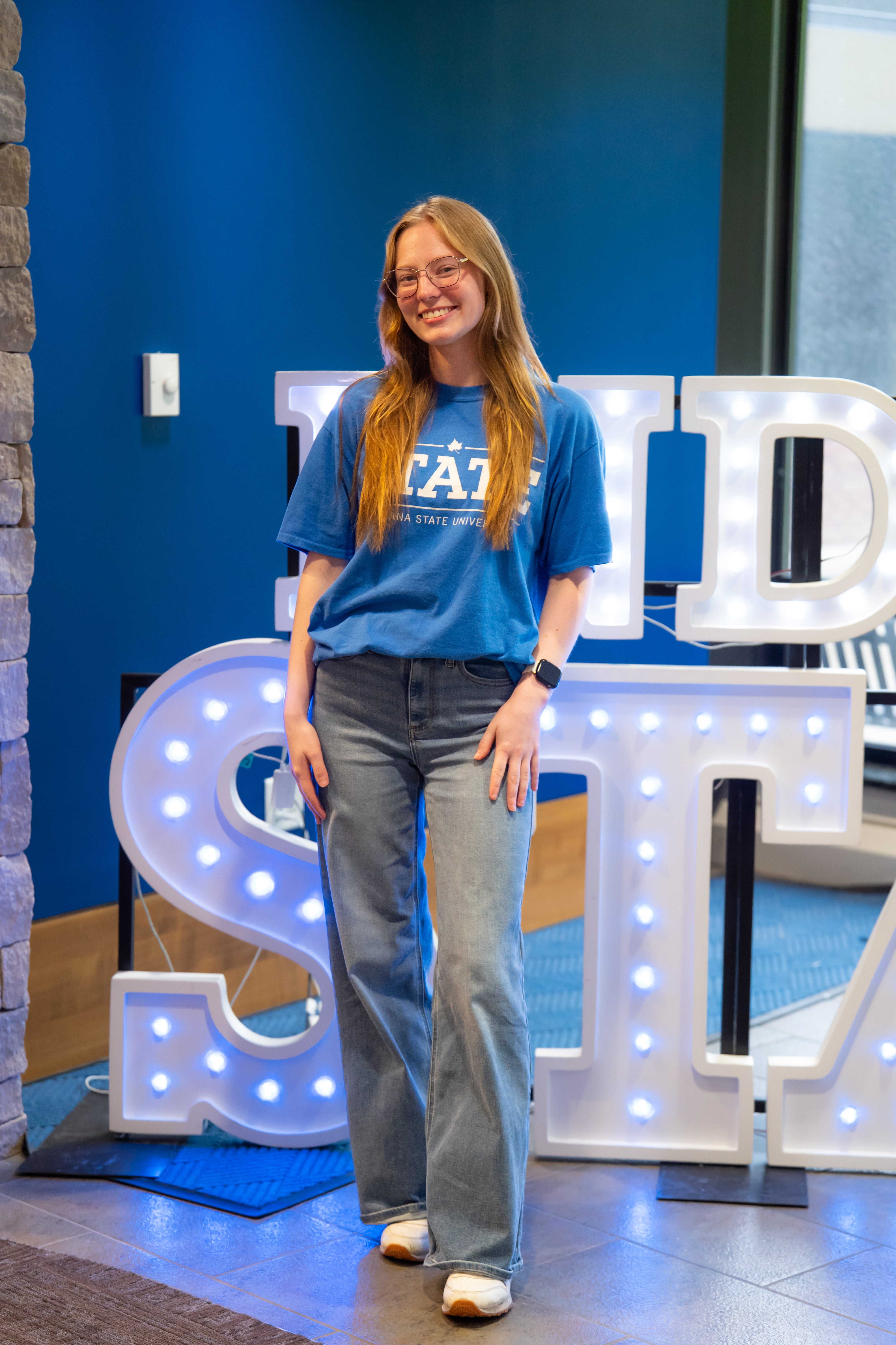 A smiling person with long blonde hair and glasses stands indoors beside illuminated “Indiana State” display letters. They are wearing a bright blue Indiana State University T-shirt, light blue wide-leg jeans, white sneakers, and a smartwatch. Blue walls, a stone wall edge, and lit signage are visible in the background.
