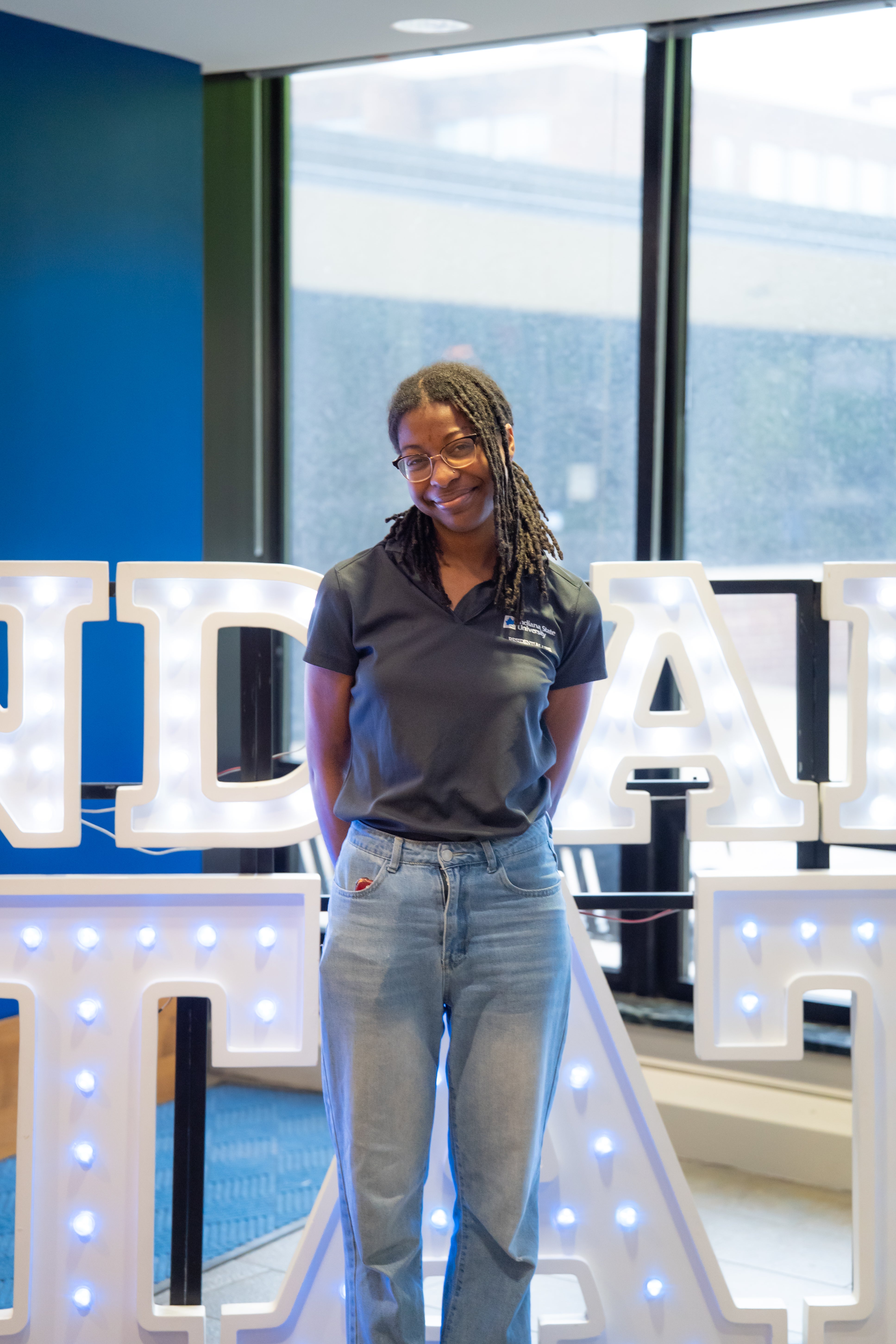 A smiling person with shoulder-length locs and glasses stands indoors in front of illuminated “Indiana State” display letters and large windows. They are wearing a black Indiana State polo shirt and light blue jeans, with their hands behind their back. Blue walls and lit signage are visible in the background.
