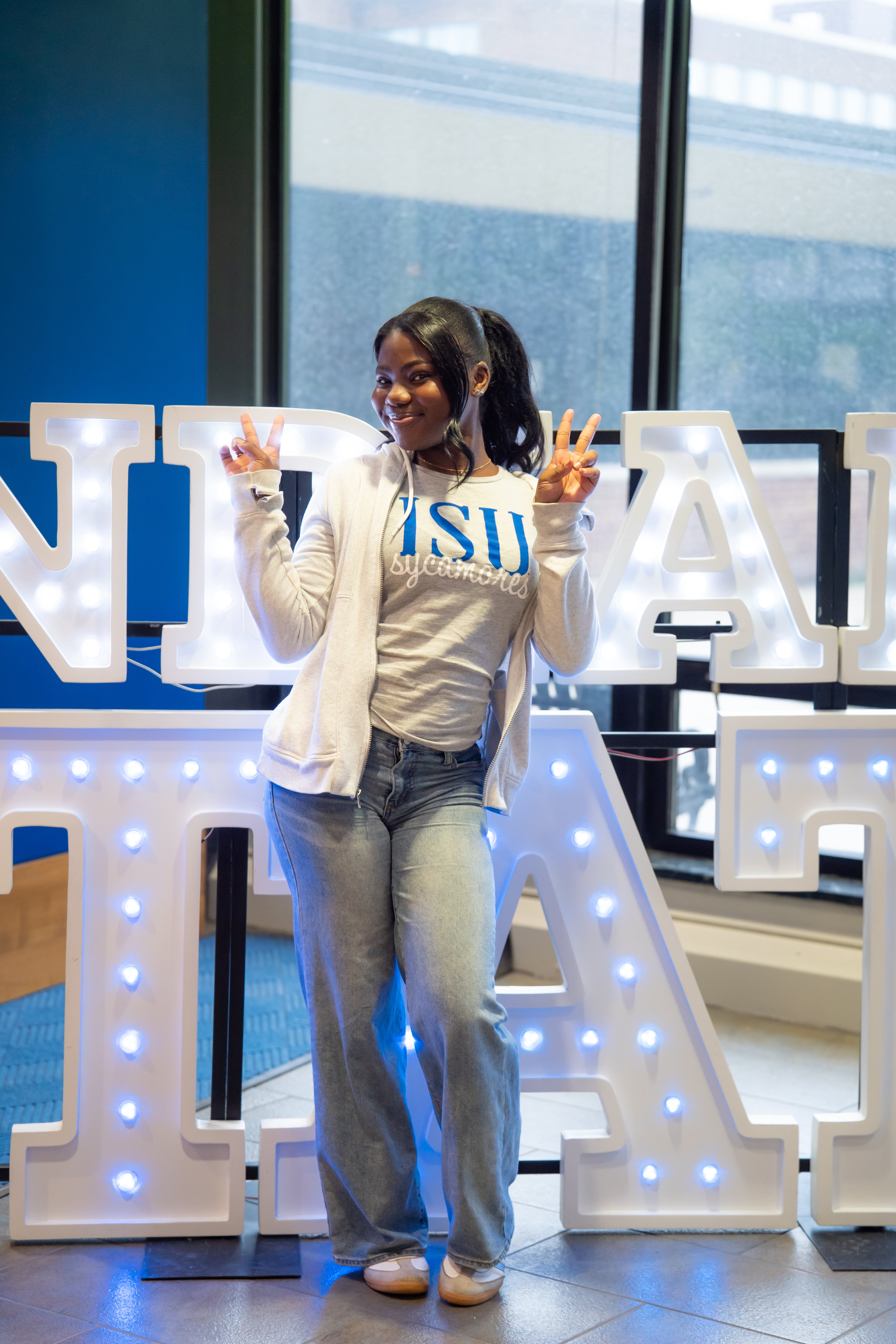 A smiling person with long dark hair in a ponytail poses indoors in front of illuminated “Indiana State” display letters and large windows, holding up peace signs with both hands. They are wearing a light gray ISU Sycamores T-shirt, a light zip-up hoodie, light blue jeans, and white shoes. Blue walls and lit signage are visible in the background.