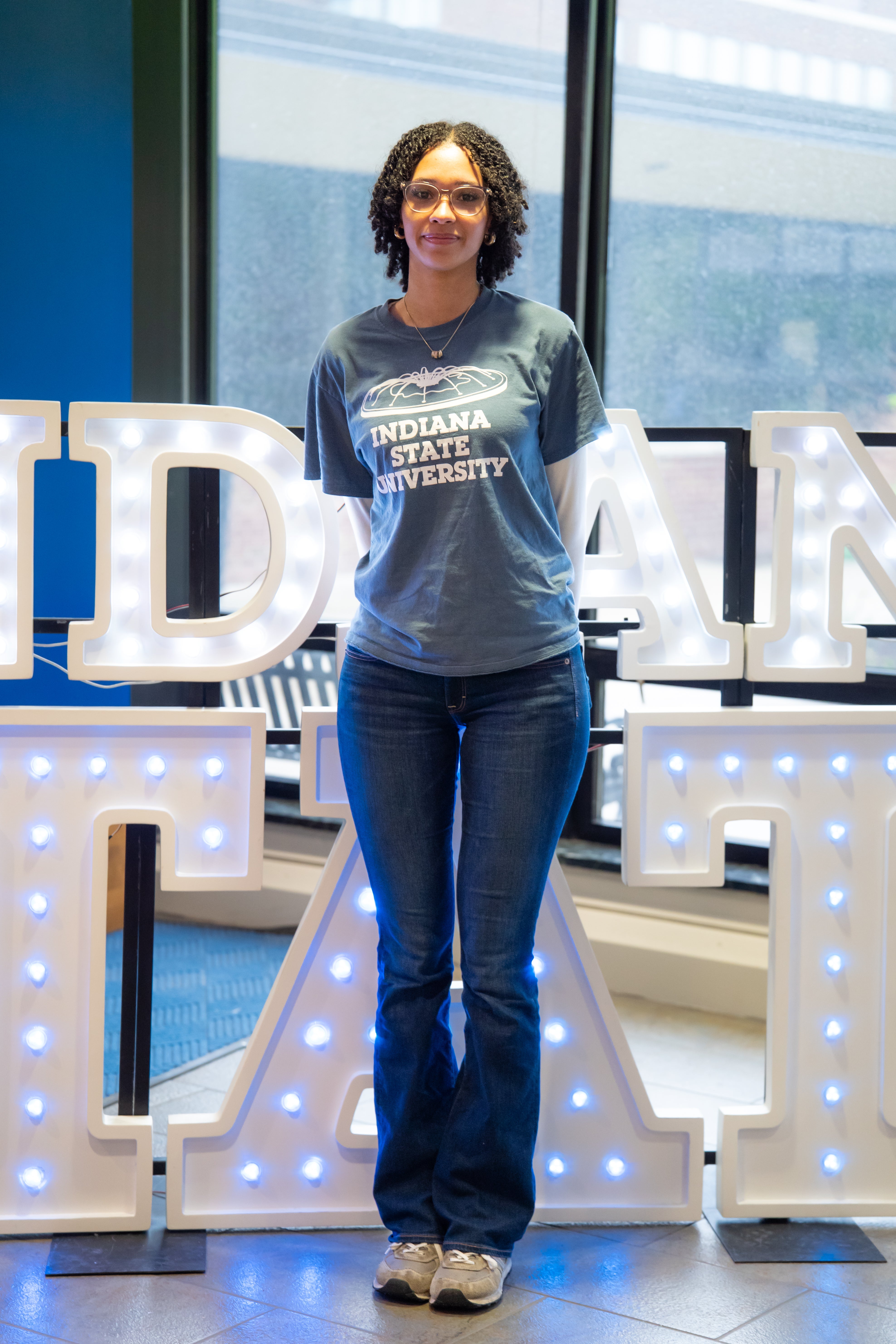 A person with short curly hair and glasses stands indoors in front of illuminated “Indiana State” display letters and large windows. They are wearing a gray Indiana State University T-shirt layered over a white long-sleeve shirt, dark blue jeans, and gray sneakers, with their hands behind their back. Blue walls and lit signage are visible in the background.