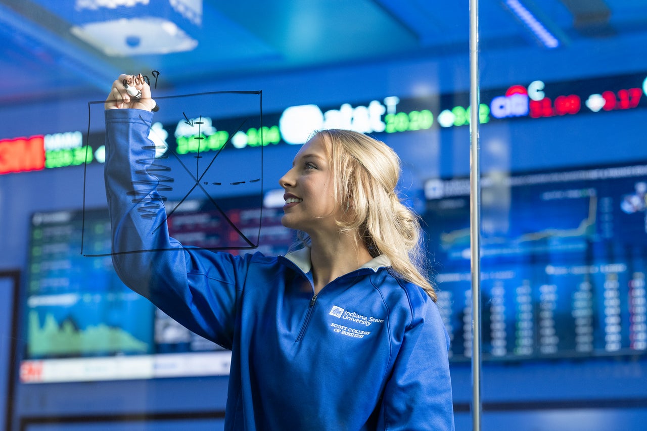 White female with blonde hair wearing a blue Indiana State University Scott College zip-up stands in front of large digital stock market displays with ticker symbols and charts, holding a marker and writing on the wall.