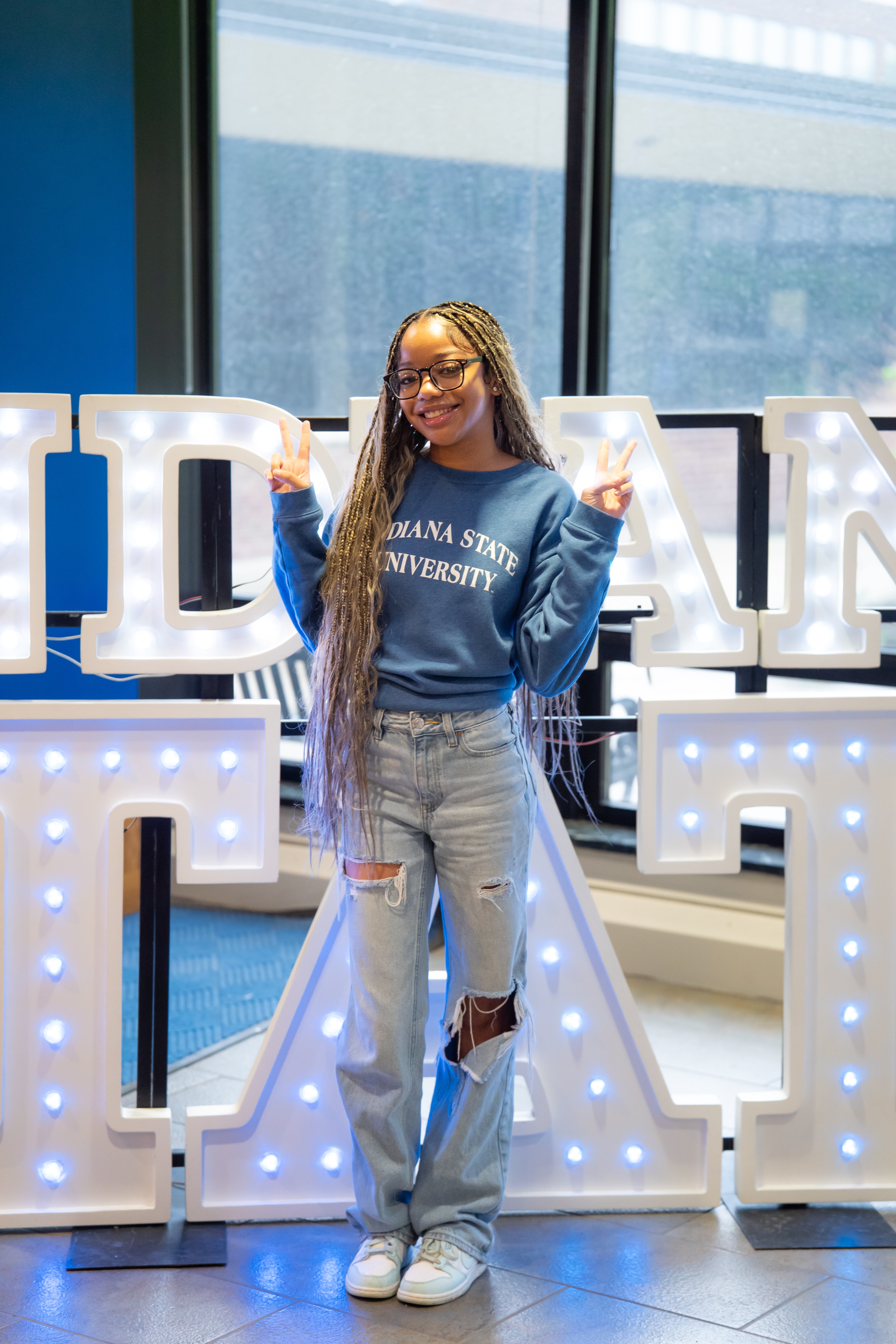 A smiling person with very long braided hair and glasses stands indoors in front of illuminated “Indiana State” display letters and large windows, holding up peace signs with both hands. They are wearing a blue Indiana State University sweatshirt, ripped light blue jeans, and white sneakers. Blue walls and lit signage are visible in the background.