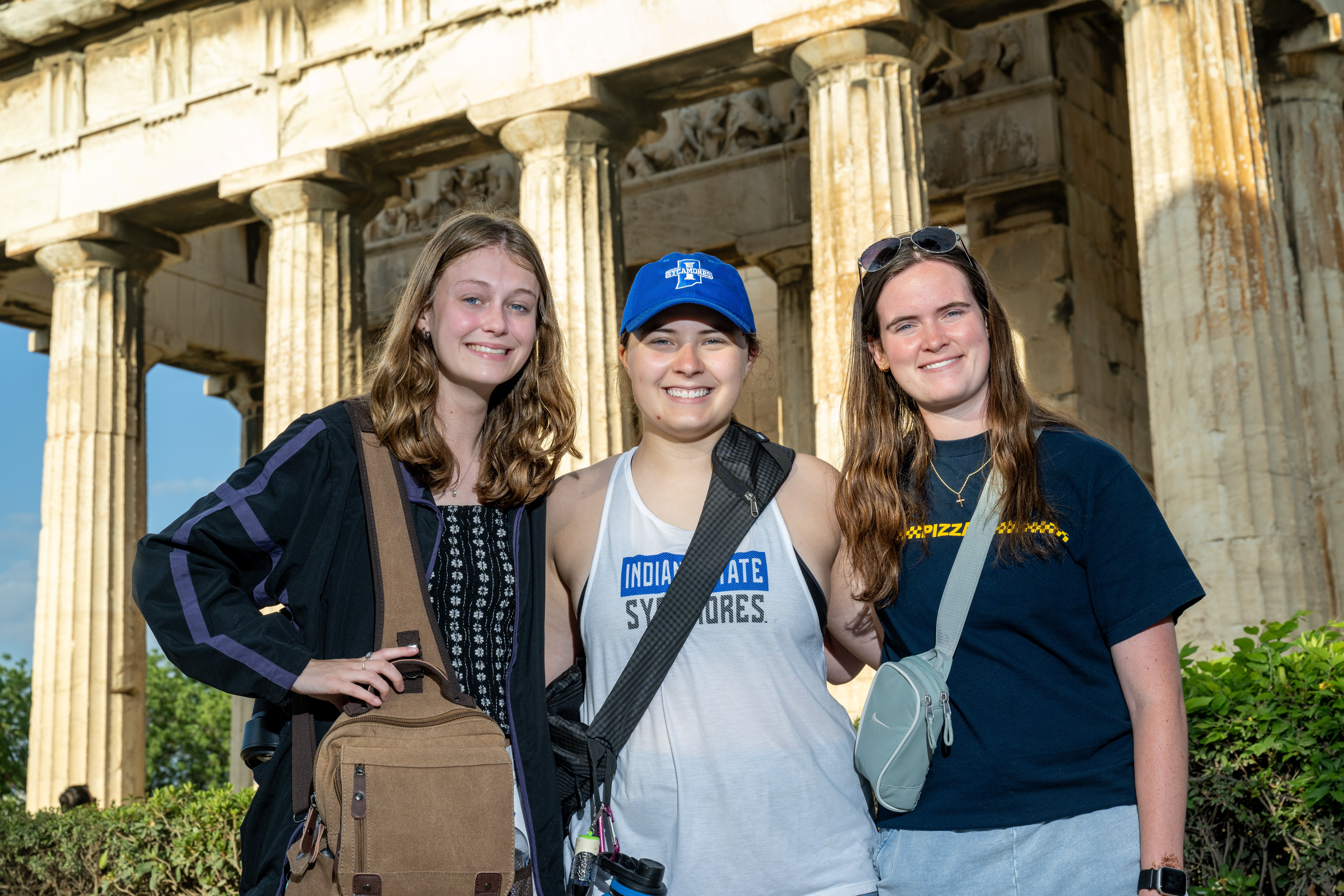 Three women stand together in front of a historic Greek stone building with tall columns, wearing casual clothing and carrying small bags, with the central person wearing an Indiana State Sycamores shirt and cap. Sunlight illuminates the structure behind them.