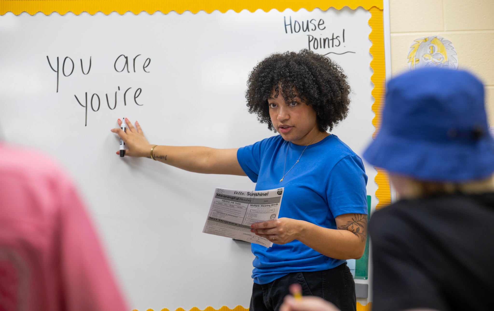 A student teacher stands at a whiteboard, pointing to the words "you are" and "you're" written on it. They hold a paper titled "Worksheet" and are wearing a blue shirt. The board is bordered with yellow paper, and "House Points!" is written in the corner. Two younger children are partially visible in the foreground.