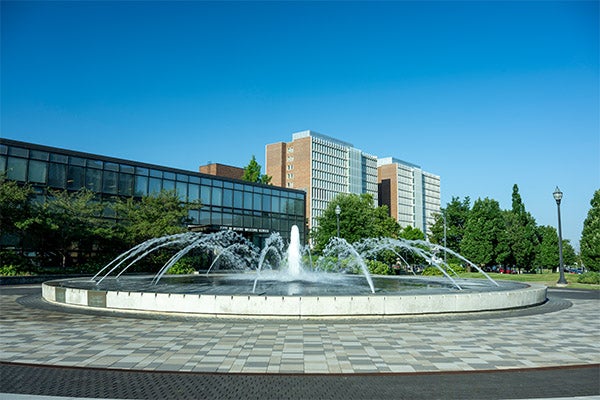 Circular fountain with multiple arcing water jets centered in a paved plaza on the Indiana State University campus. Low, modern academic buildings and taller residence hall-style buildings stand behind the fountain, with green trees and a clear blue sky in the background.