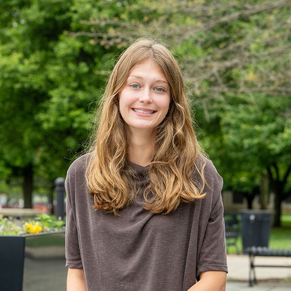 Smiling young woman with long hair sits on an outdoor campus walkway near a large planter, with trees, benches, and a campus building in the background.