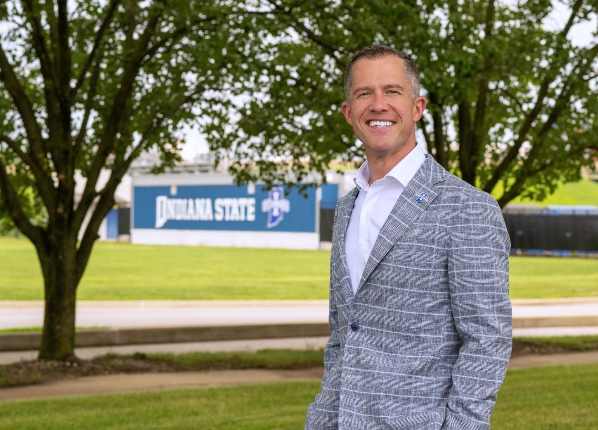 A person in a gray plaid suit stands smiling outdoors with trees and grass in the background. A building with signage that reads "Indiana State" is visible in the distance.