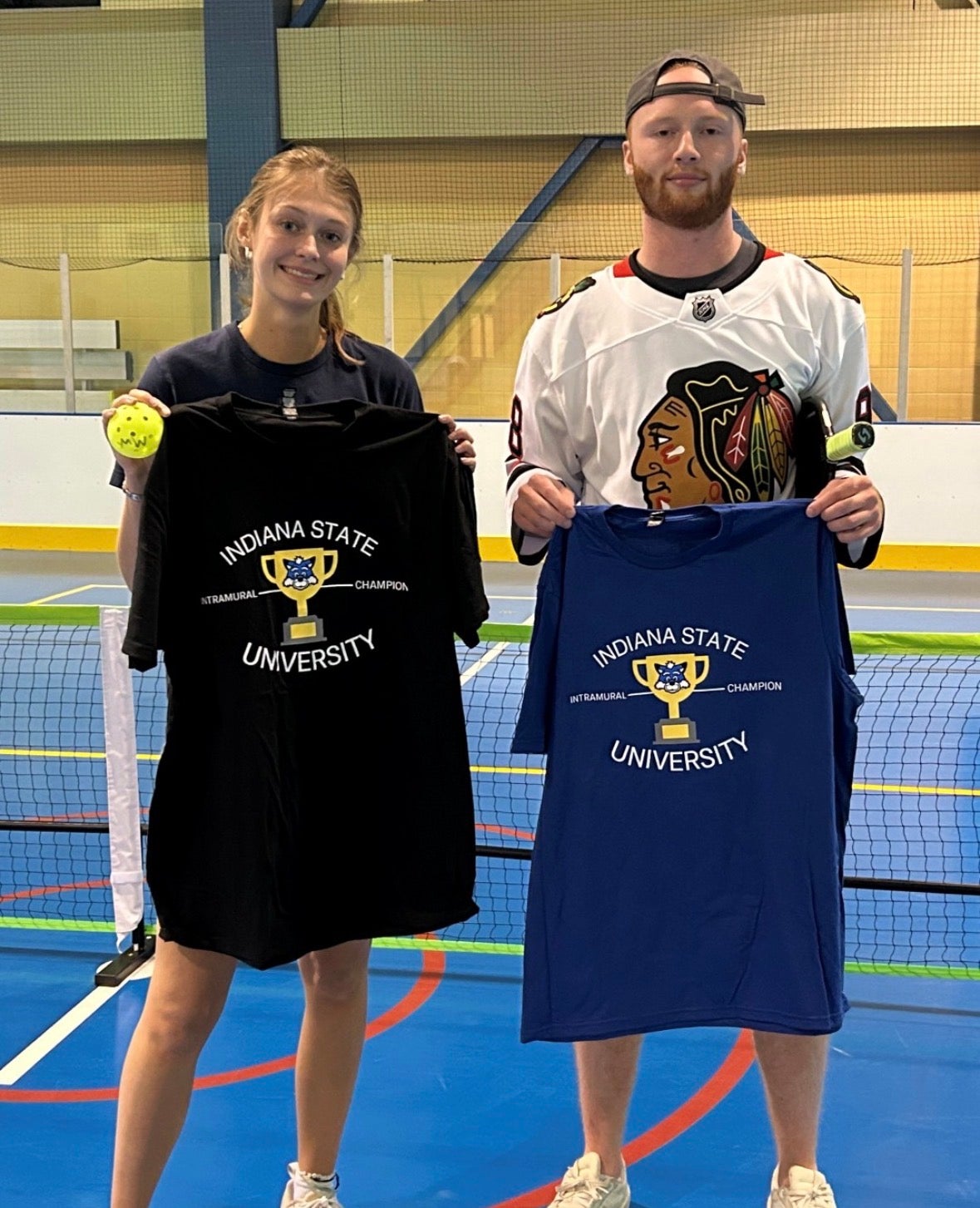 Two people stand on an indoor sports court holding Indiana State University Intramural Champion T-shirts, one black and one blue, with a tennis net and court lines visible in the background.