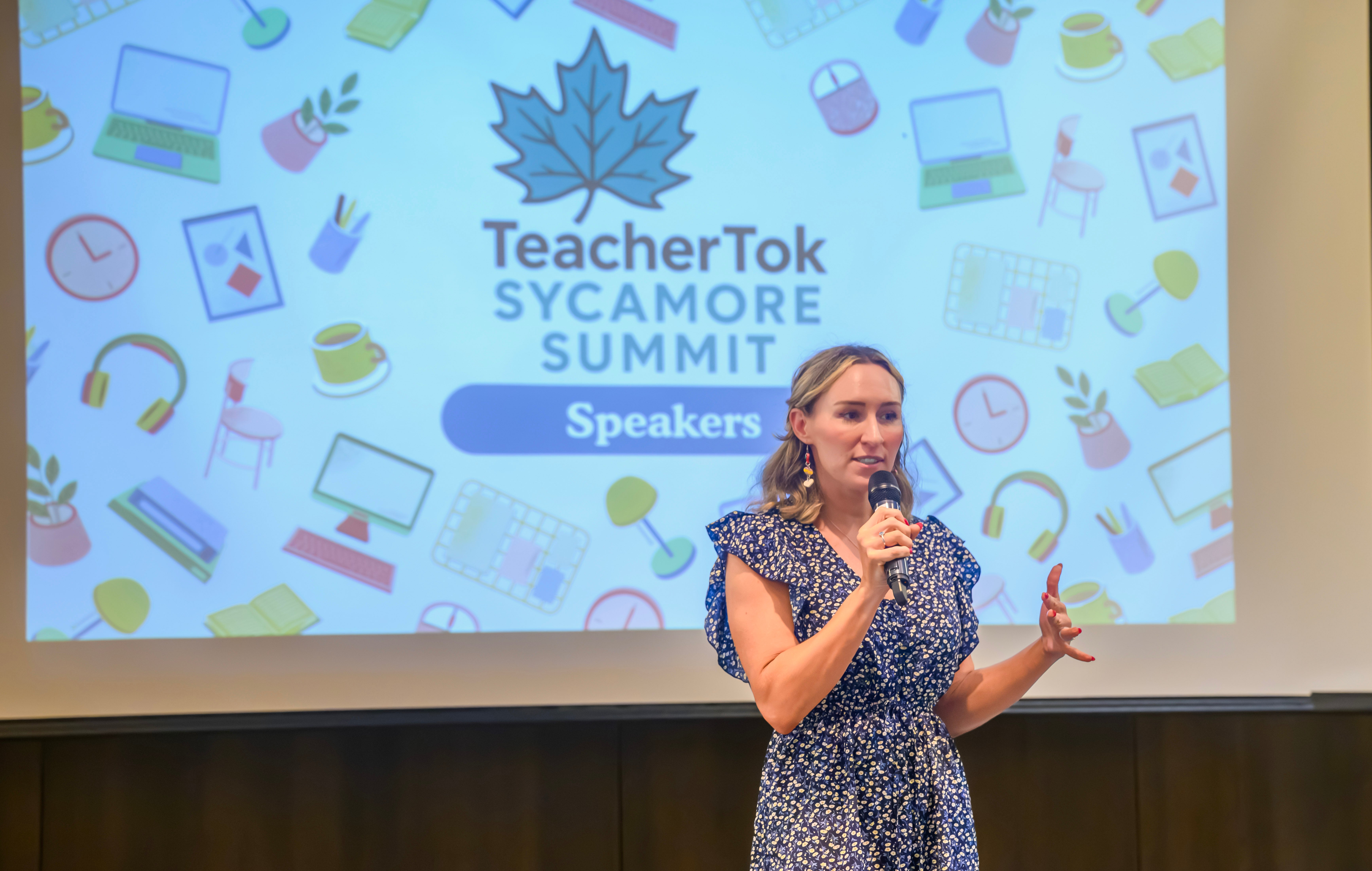 An individual stands holding a microphone, speaking in front of a projector screen displaying graphics and the text "TeacherTok Sycamore Summit." The screen also has the word "Speakers" highlighted.