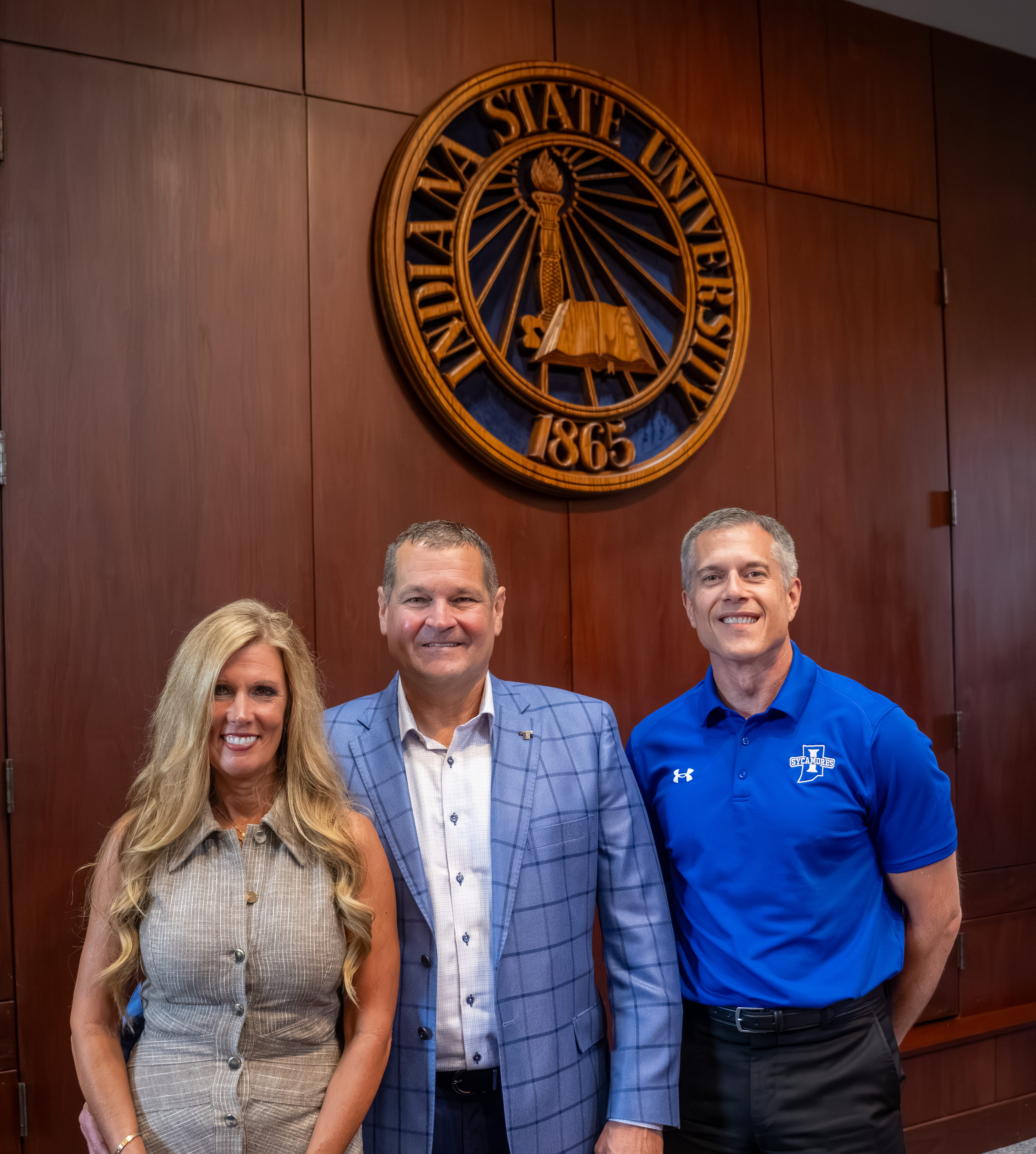 Three people pose beneath the Indiana State University seal in a campus boardroom.