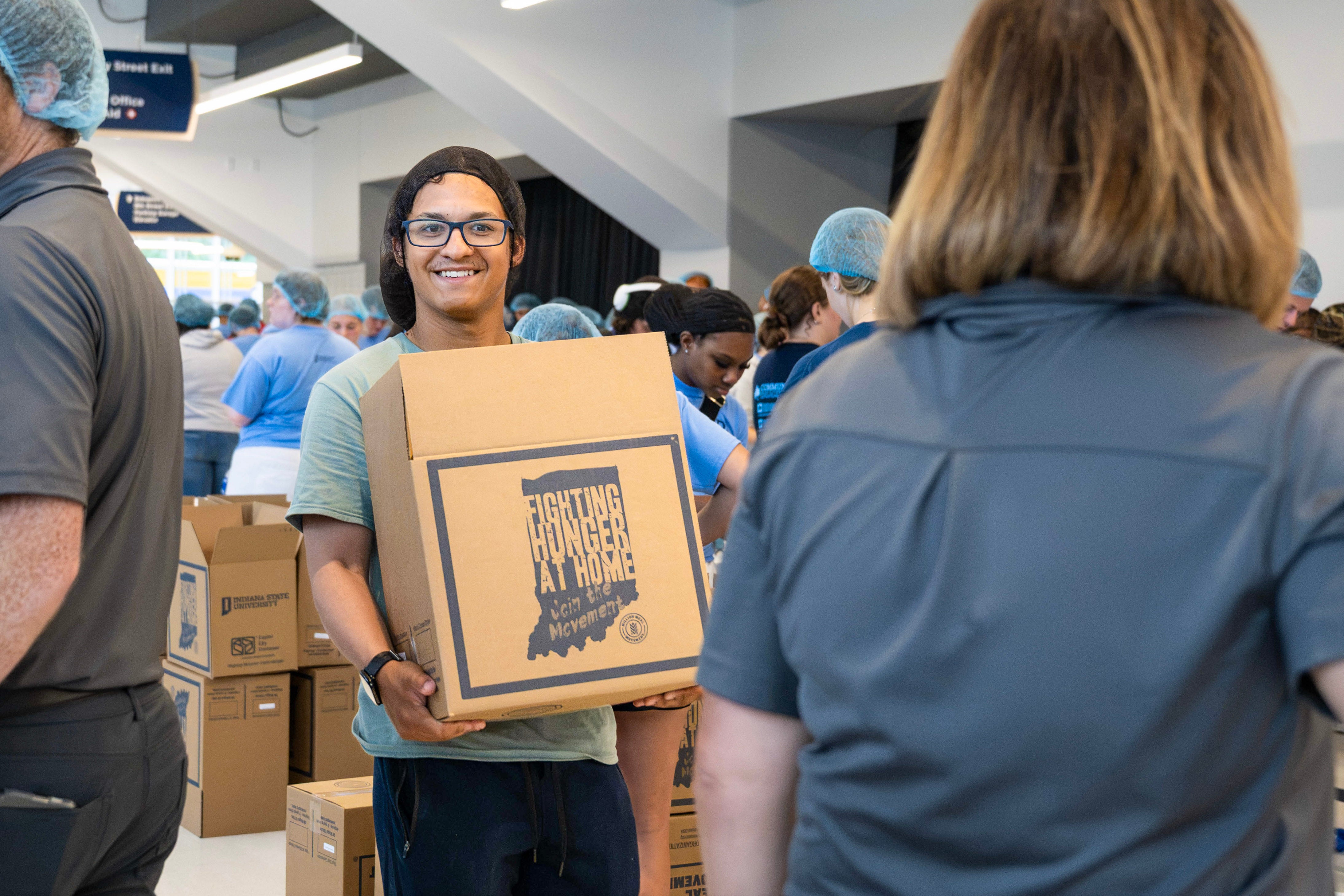 A person carrying a box labeled "Fighting Hunger at Home" at a community event. People in the background are wearing hair nets and sorting boxes.
