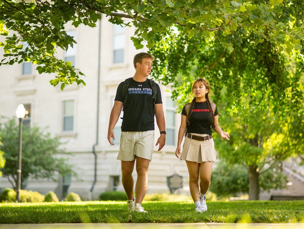 Two students walk across a grassy campus area beneath large shade trees. One student carries a backpack, and a campus building is visible in the background.