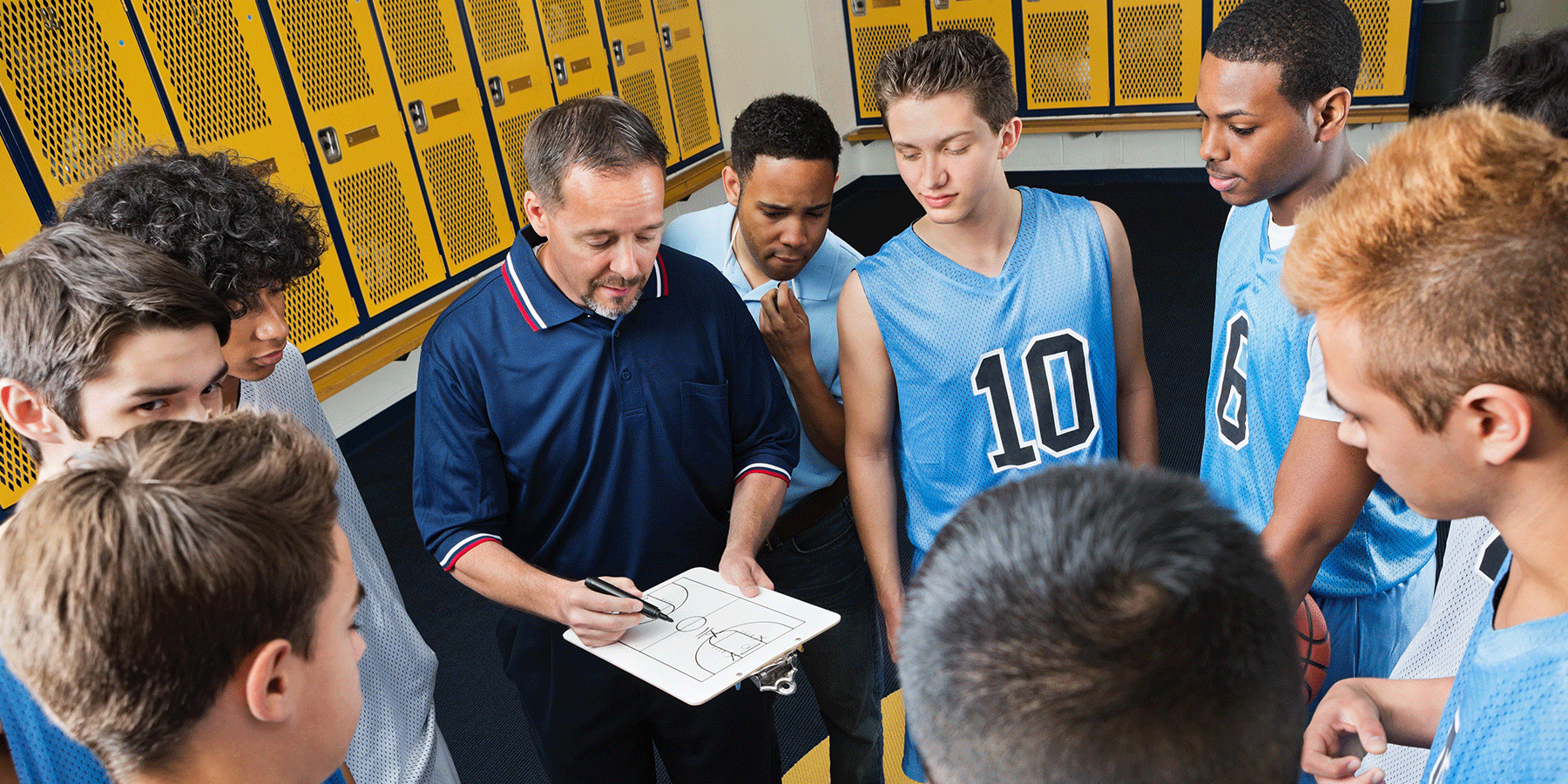 Coach holding a clipboard with a basketball court diagram while standing in a locker room surrounded by players in practice jerseys. Yellow lockers line the walls as the group gathers closely to review the coach’s instructions.