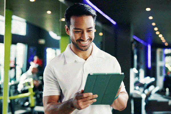 Person standing in a fitness center holding a tablet and looking at the screen. Exercise equipment, mirrors, and bright green columns are visible in the background, with overhead lights reflecting throughout the gym.