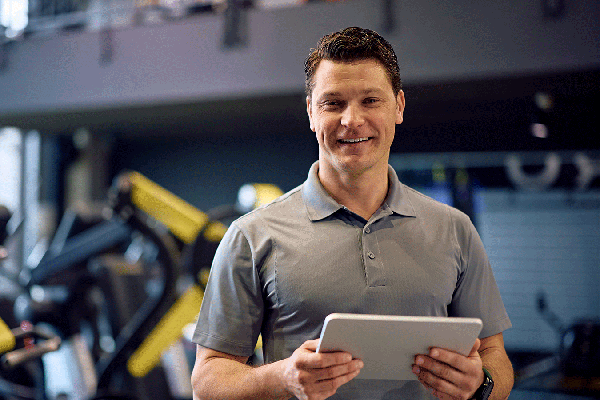 Smiling man in a gray polo shirt holding a tablet while standing in a fitness center. Exercise equipment with yellow accents is visible in the blurred background, and natural light enters from windows on the left.